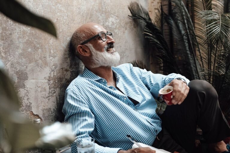 An older man with a white beard laughing as he writes in a notebook at a café table